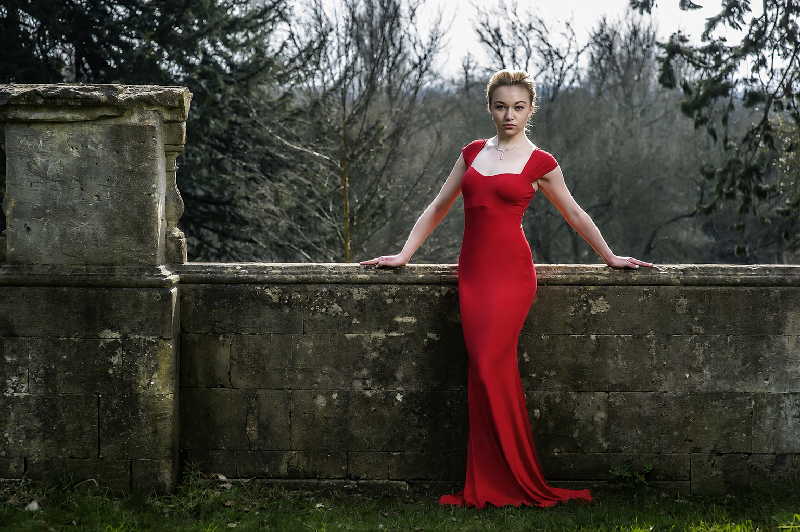 A woman in a long red dress stands outdoors against an old stone wall, with bare trees and muted greenery creating a rustic, natural backdrop. The vivid red dress contrasts with the weathered stone, highlighting a classic portrait photography style focused on colour contrast, texture, and environmental storytelling.